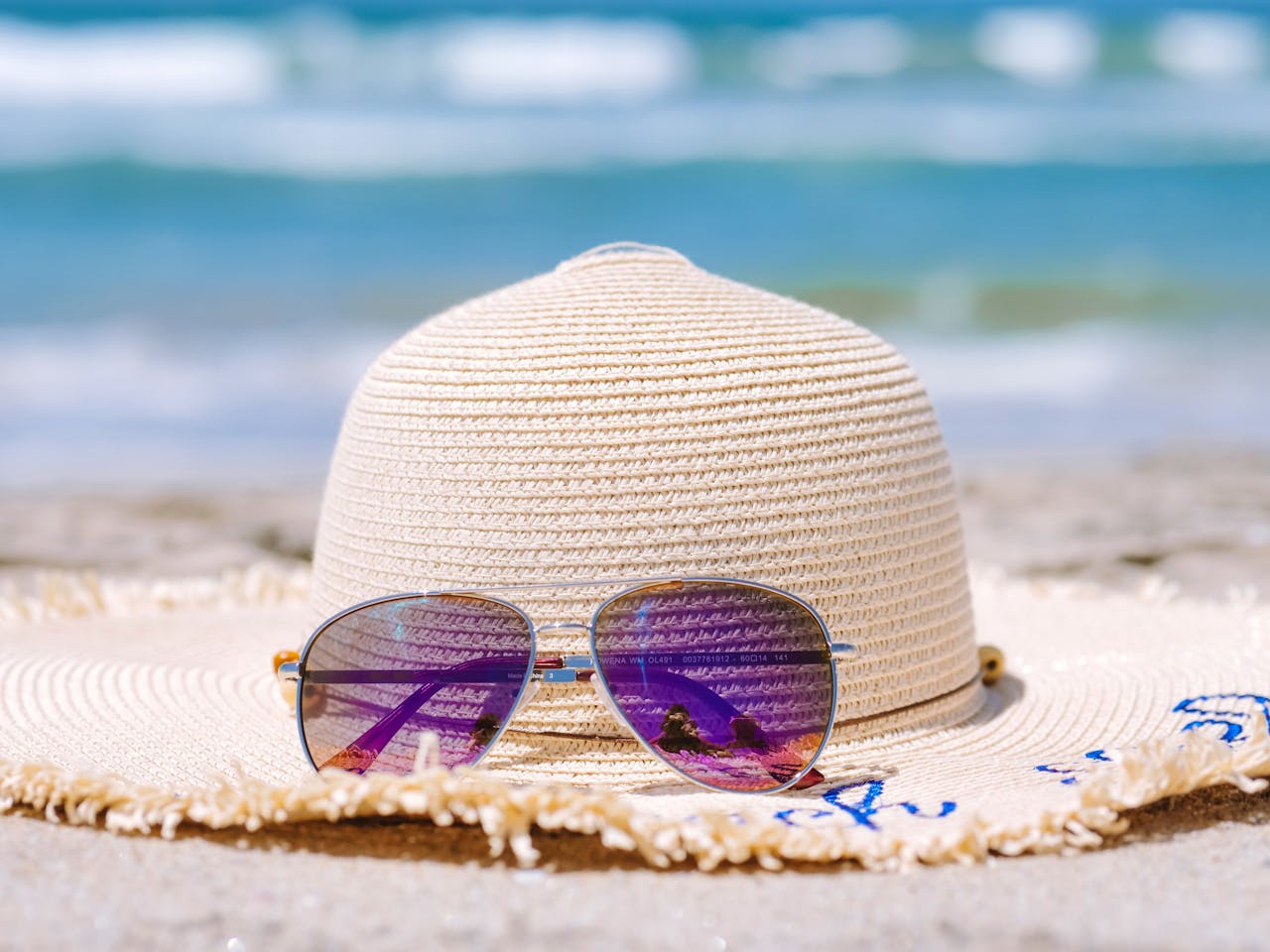 A sun hat and sunglasses rest on a beach, reflecting a beautiful seascape.