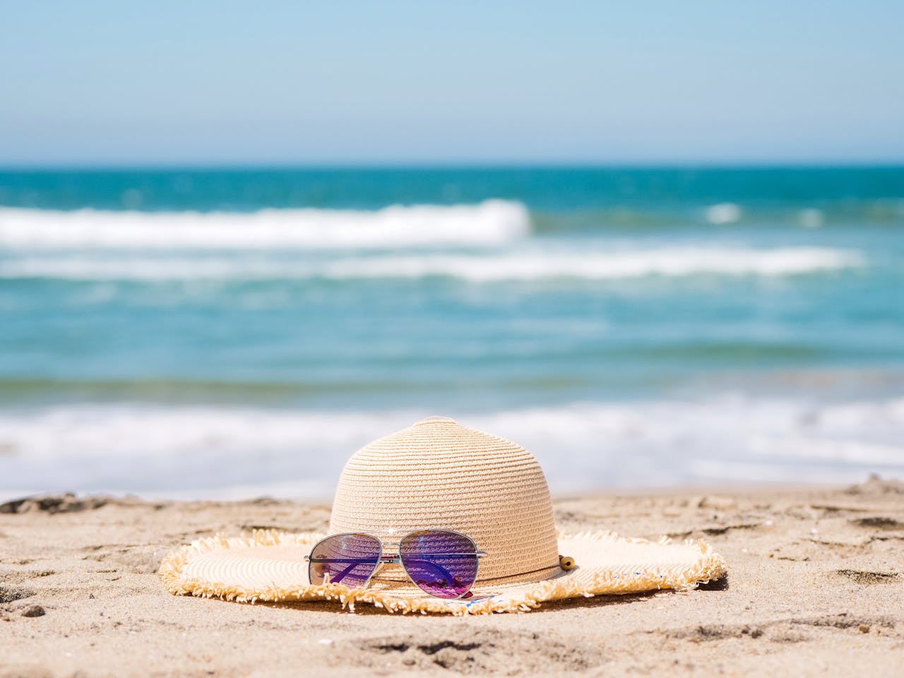 A straw hat and sunglasses resting on a sandy beach with blue waves in the background.